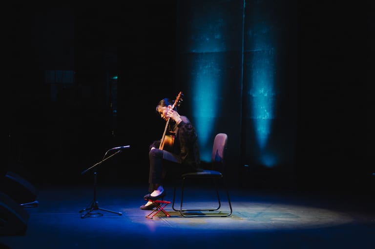 Cellist performs solo on stage beneath blue spotlights in a darkened theatre.