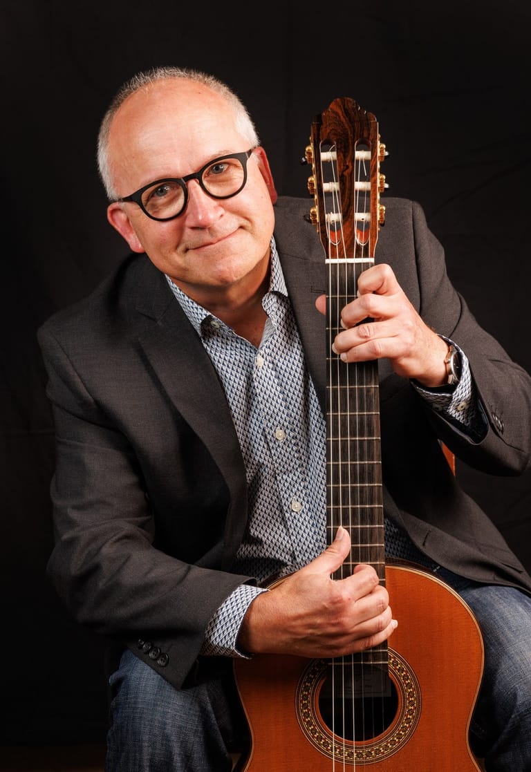 Man in glasses and blazer holding a classical guitar against a black background.