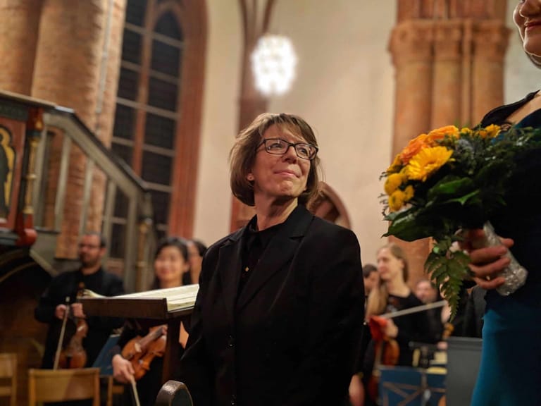 Person in glasses holding a programme stands in a classical venue with orchestra musicians and yellow flowers visible in background.