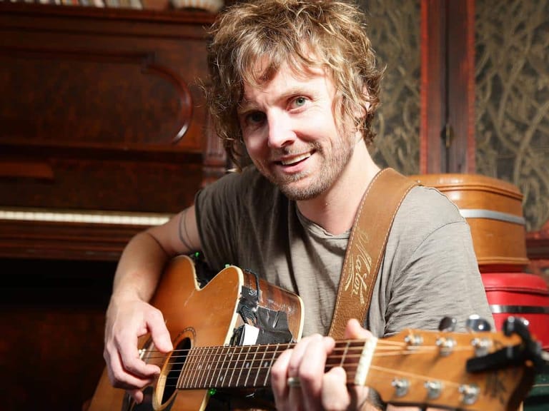 Musician with shoulder-length hair smiling while playing an acoustic guitar in a warm, richly decorated interior space.
