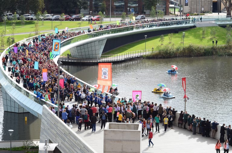 Large crowds gather along a curved waterside promenade beside a modern bridge and canal.