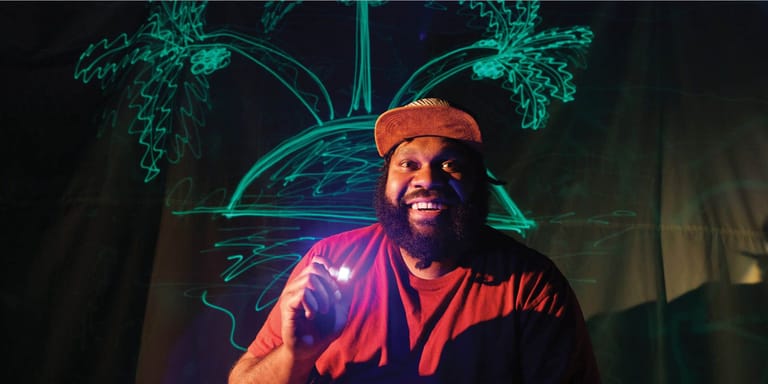 Man in orange shirt surrounded by glowing teal plant projections and purple stage lighting.
