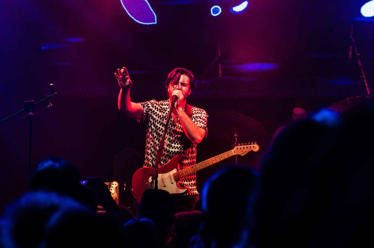 Performer in studded gold jacket plays electric bass guitar on stage under purple and blue stage lighting.