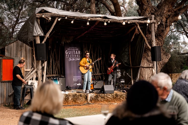 Two musicians perform on a rustic outdoor stage beneath strung lights, surrounded by seated audience members.