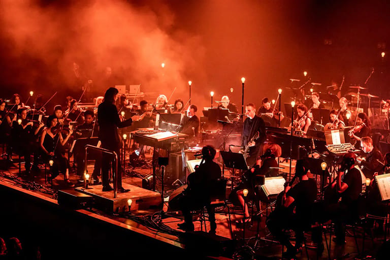 Large orchestra and choir perform together on stage bathed in warm orange and red lighting.