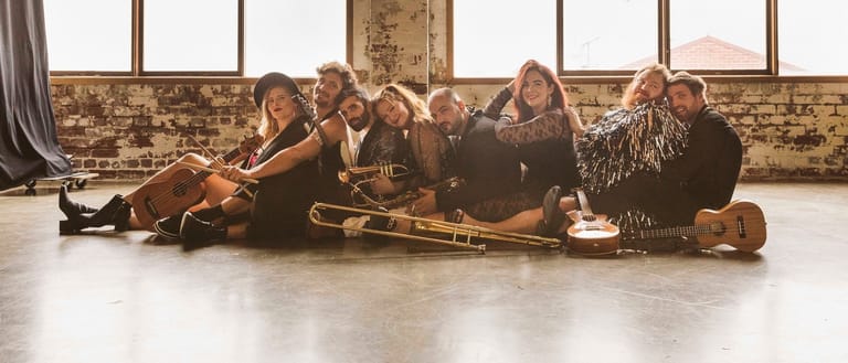 Six musicians sit cross-legged on a polished floor with brass instruments in a brick studio space.