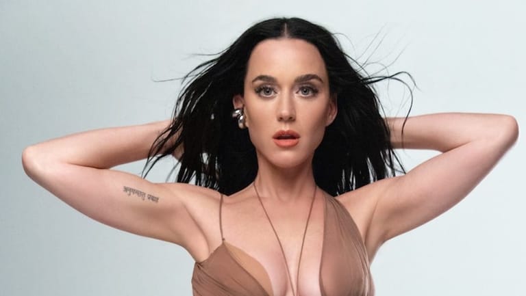 Woman with long black hair and visible tattoo on arm, hands behind head against neutral grey background.
