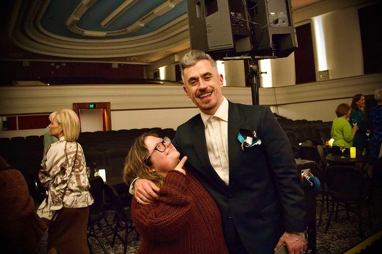 Man in dark suit and white shirt embraces woman in burgundy top at an elegant theatre venue.