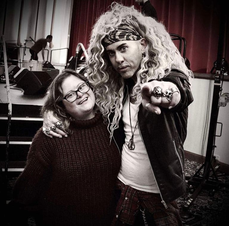 Two people pose together backstage, one wearing a long curly blonde wig and headband, against a burgundy curtain with stage equipment visible.