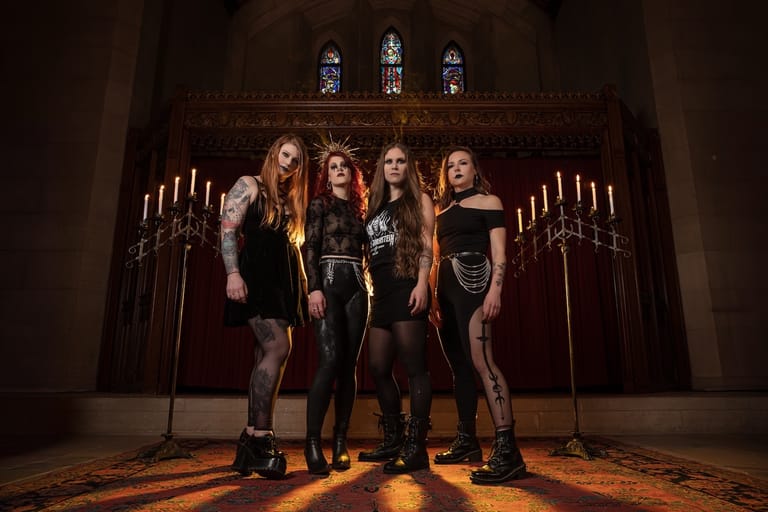 Four musicians stand together in a gothic chapel setting lit by candlelight and orange floor lighting.