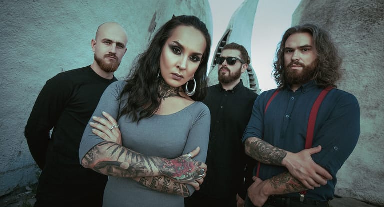 Four band members pose against a textured wall, with the female vocalist in front wearing a grey top and dark makeup.