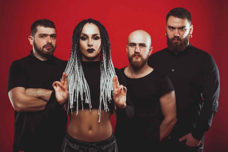 Four band members posed against a red background, with the female vocalist in the centre wearing white braided hair and black clothing.