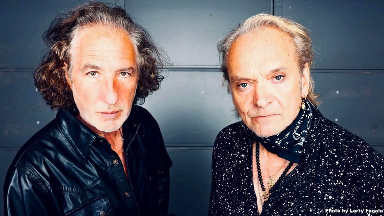 Two musicians with long hair pose against a grey wall, one wearing a black jacket, the other in a sparkly black shirt.
