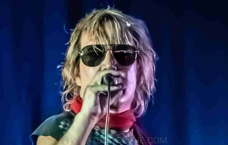 Performer wearing dark sunglasses singing into microphone under blue and pink stage lighting.