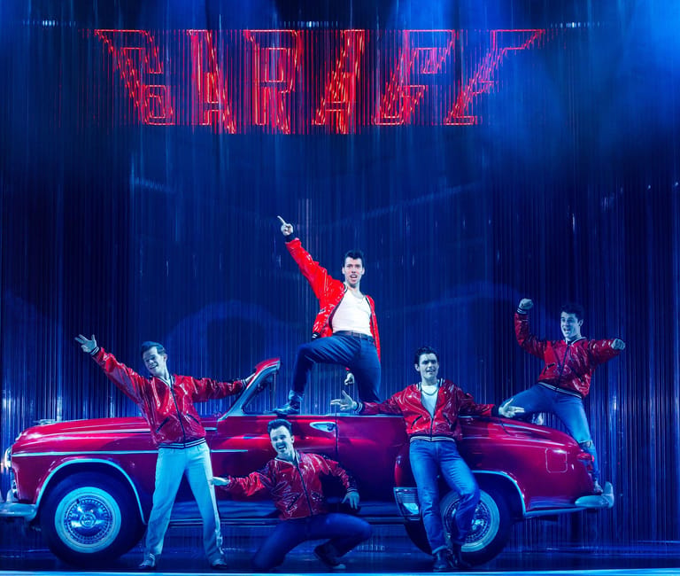 Performers in red jackets and jeans dance on and around a vintage pink car beneath neon "GREASY" signage on a blue-lit stage.