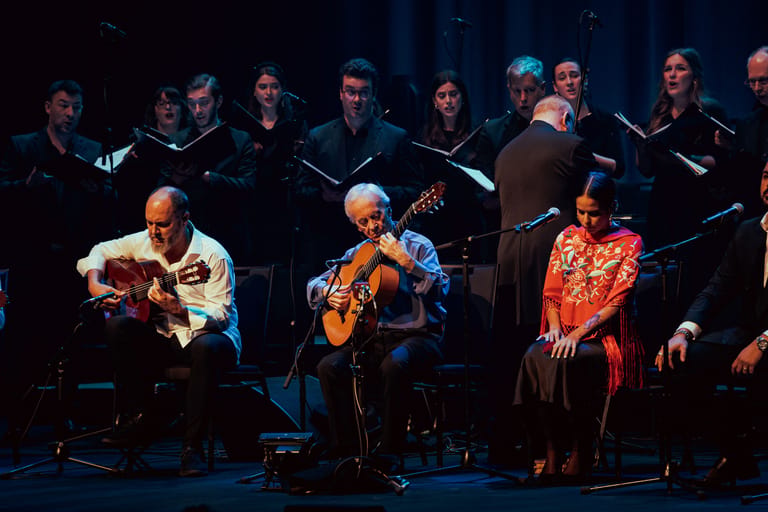 Three guitarists perform on stage with a choir ensemble seated behind them under blue theatrical lighting.