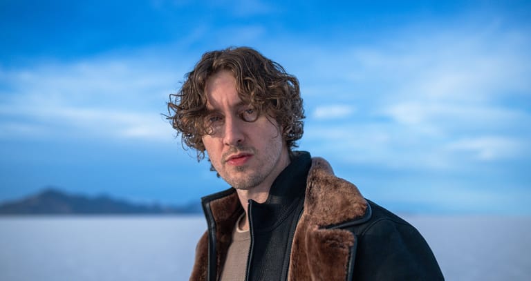 Young man with curly hair wearing a brown and black jacket against a misty mountain landscape.