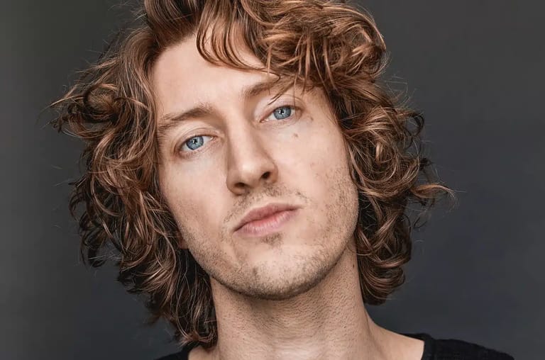 Person with shoulder-length curly brown hair and blue eyes against a grey background.