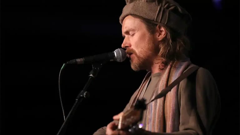 Musician singing into a microphone while playing guitar, wearing a brown cap and shirt on a dark stage.