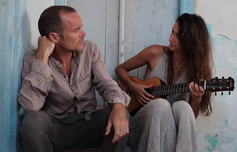 Two musicians sit against a weathered blue wall, one holding an acoustic guitar in an intimate performance setting.