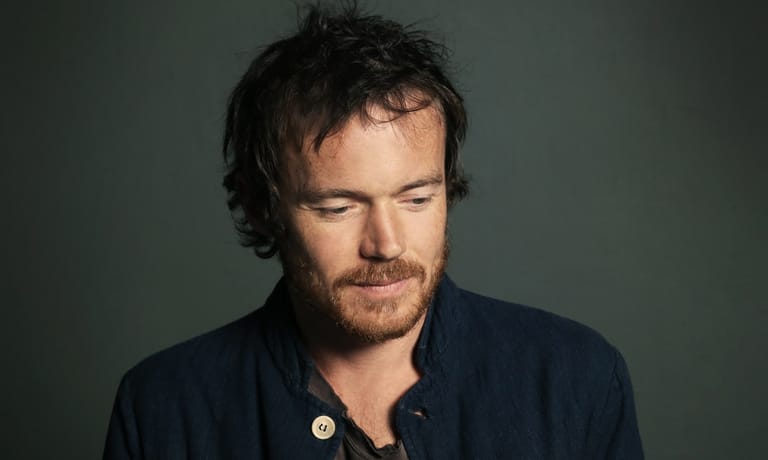 Man with shoulder-length dark hair wearing a navy shirt against a grey-green background.