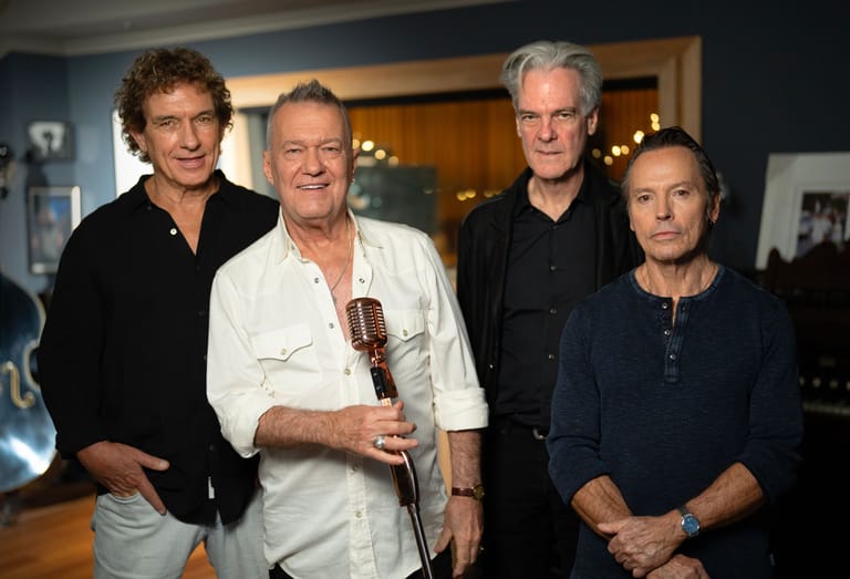 Four musicians pose together indoors, with the second from left holding a brass instrument.