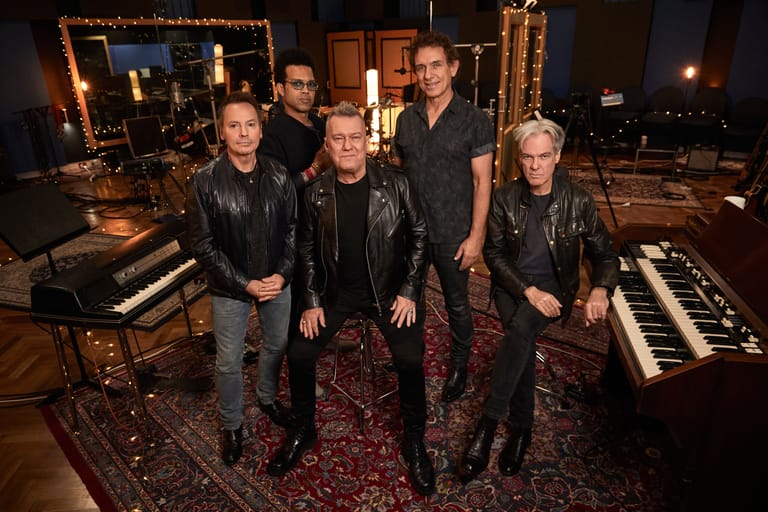 Five musicians in black leather jackets stand in a warm-lit studio with keyboards and stage lighting.