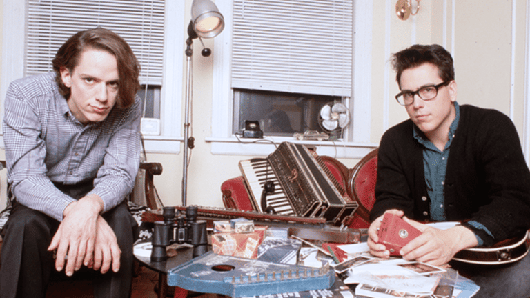 Two musicians sit at a cluttered desk with a keyboard and vinyl records in a home studio setting.