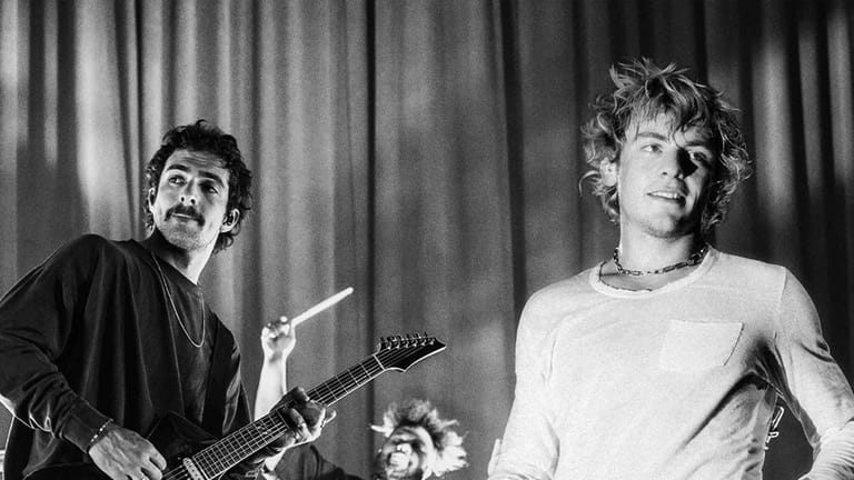 Guitarist and vocalist perform together on stage in black and white, with draped curtains behind them.