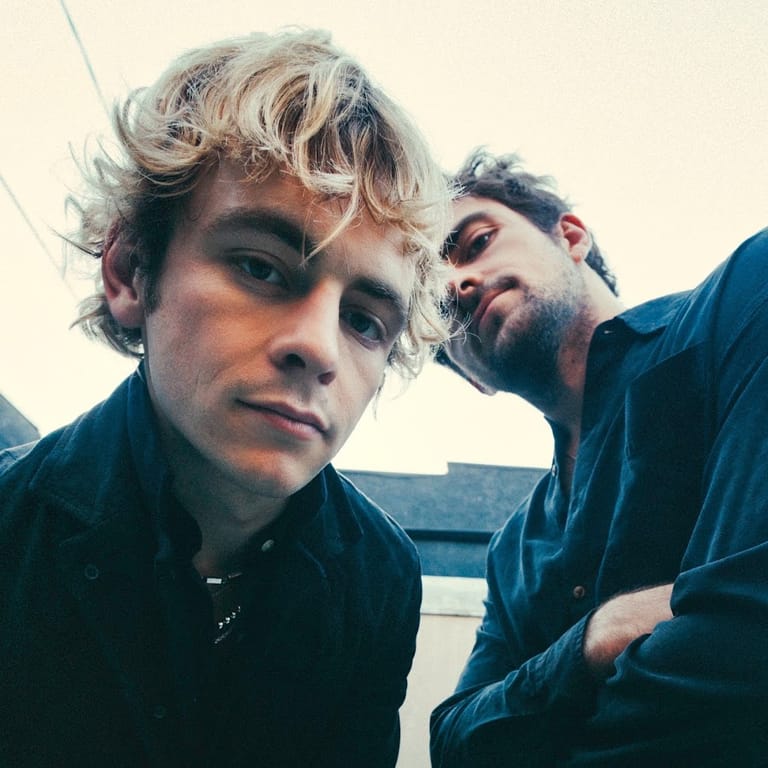 Two musicians in dark shirts pose closely together against a neutral sky backdrop.