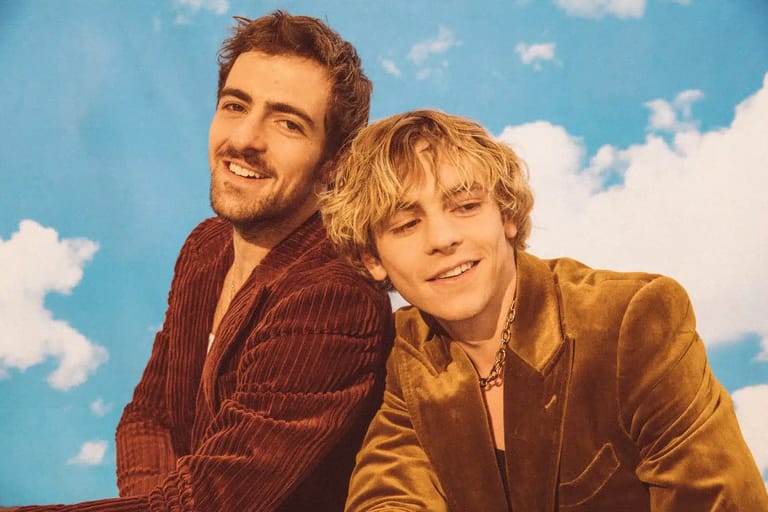 Two smiling young men pose against a bright blue sky backdrop with white clouds.