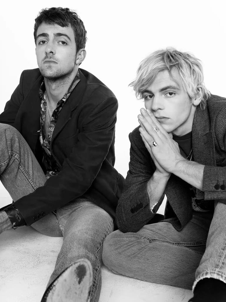 Two men in dark jackets and jeans sit against a white background in a black and white portrait photograph.