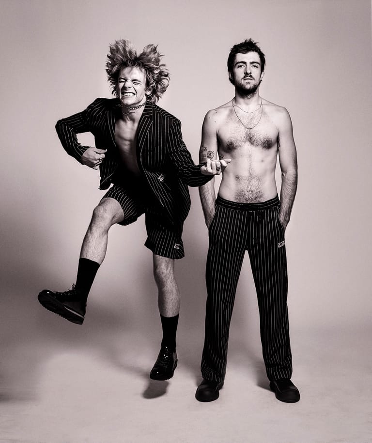 Two men pose against a neutral background; one wears a pinstriped blazer with leg raised, the other shirtless in pinstriped trousers.