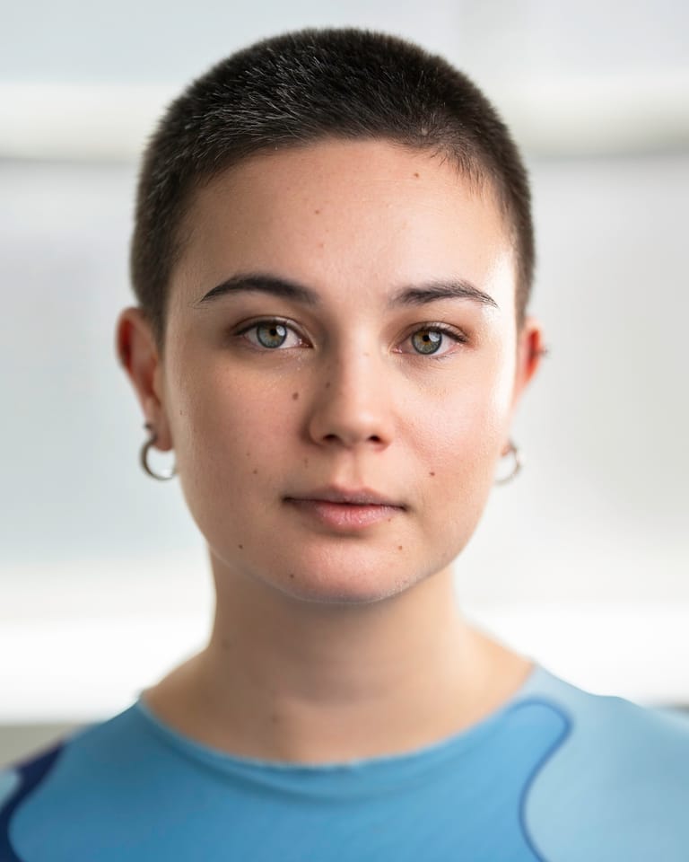 Person with short dark hair and silver ear piercings wearing a light blue shirt, neutral expression against a blurred white background.