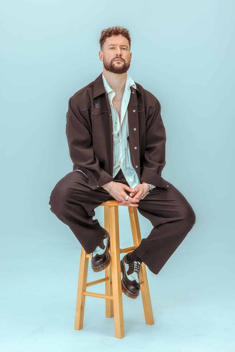 Man in brown suit seated on wooden stool against light blue background.