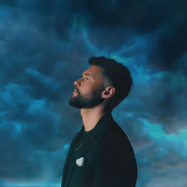 Man in profile gazing upward against a dramatic blue-toned cloudy sky backdrop.