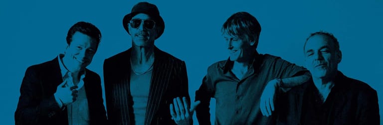 Four men in suits posed against a blue backdrop, lit by cool stage lighting.
