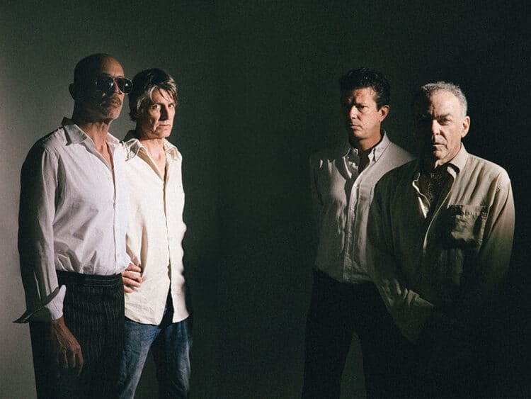 Four men in casual shirts stand against a dark background in moody stage lighting.