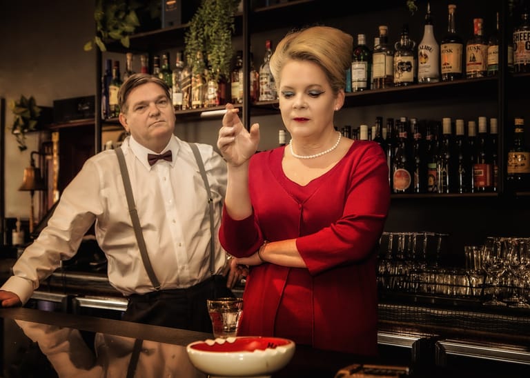 Woman in red dress and man in bow tie at a bar with bottles lining shelves behind them.