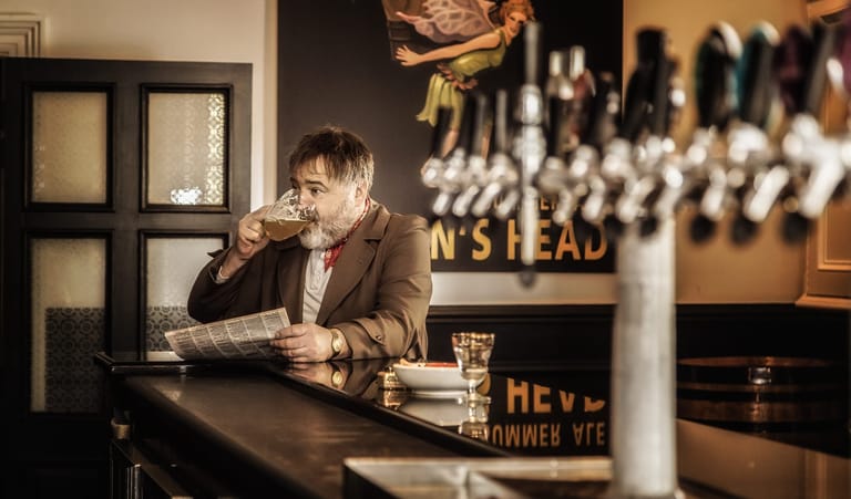 Man in brown jacket sits at bar counter, holding drink and looking pensively downward in dim interior setting.