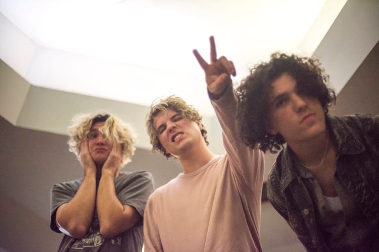 Three musicians pose together with one making a peace sign, shot from below against a bright ceiling.