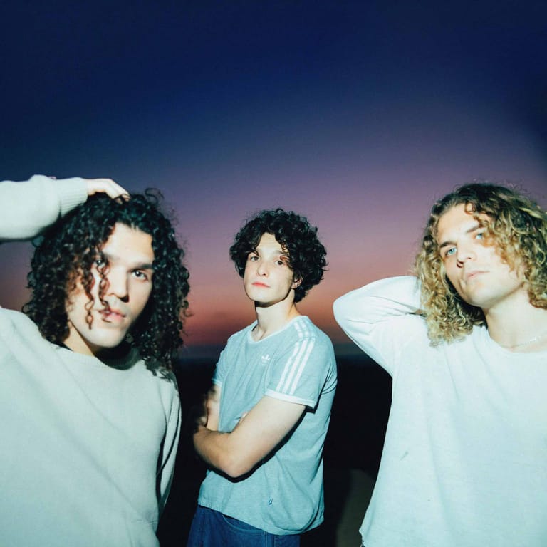 Three musicians with curly hair pose against a gradient sunset sky backdrop.