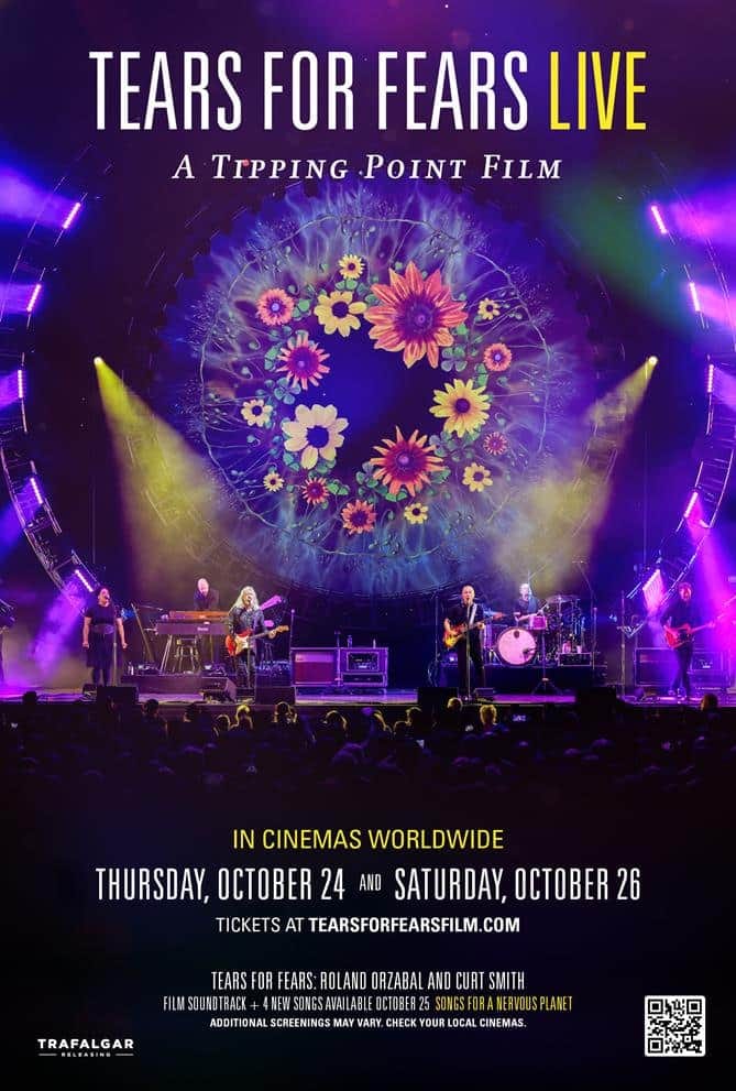Tears for Fears performing on stage beneath a large floral projection, bathed in purple and yellow stage lighting.