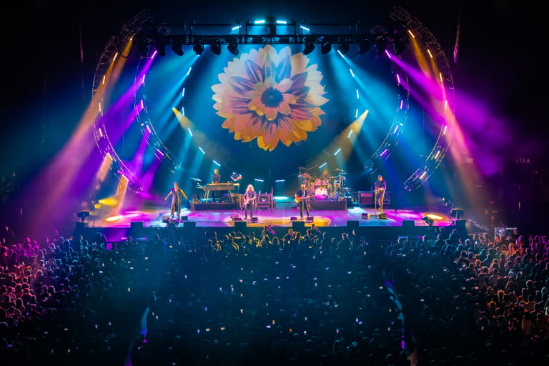 Large crowd watches a concert stage illuminated by vibrant purple and blue lighting beneath a sunflower projection.