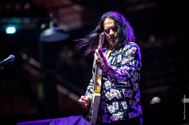 Musician with long dark hair performing with guitar on stage under purple and blue lighting.