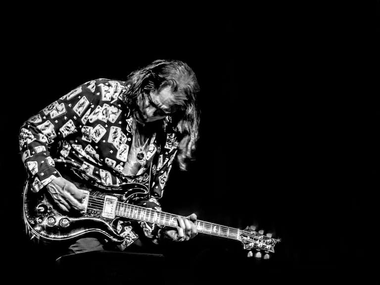Musician in patterned shirt playing electric guitar against dark background.