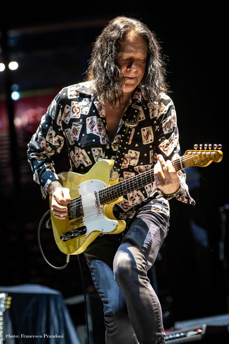Guitarist in a patterned jacket performing with a yellow electric guitar on stage.