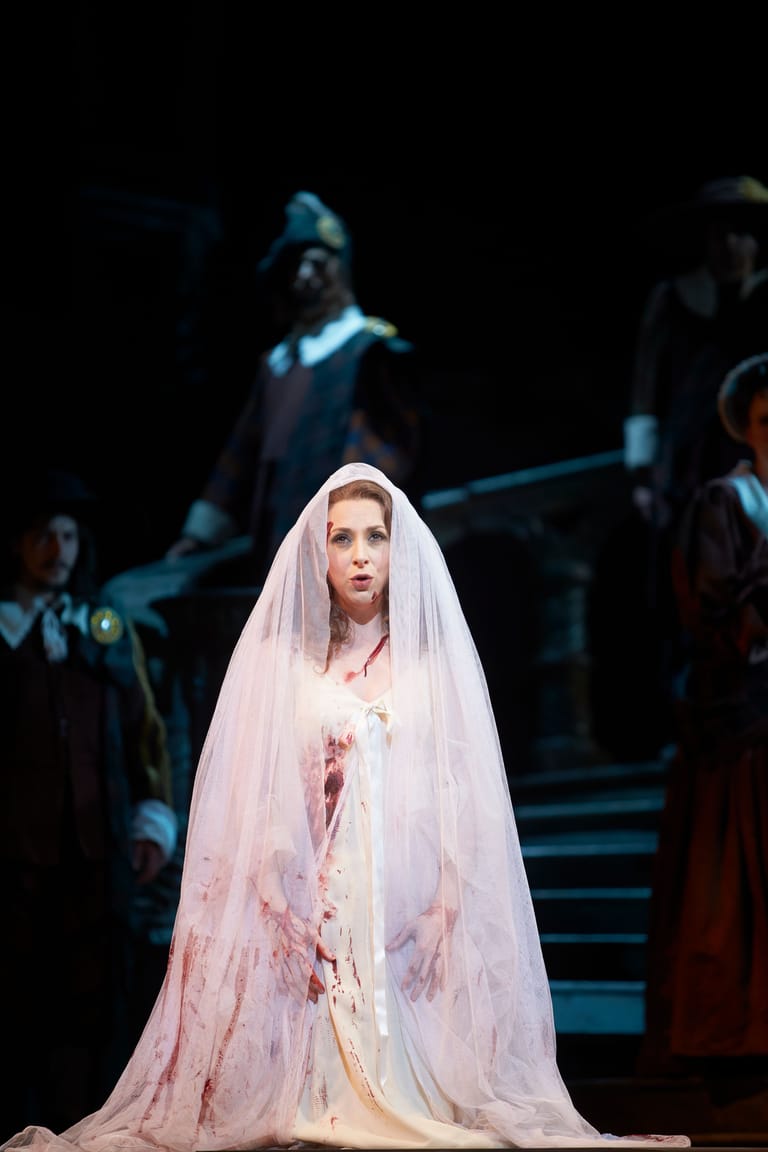 Performer in white veil and bloodstained gown stands centre stage in dramatic theatrical production.