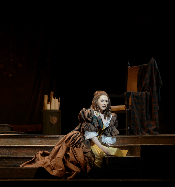 Woman in period costume sitting on wooden stage set under warm spotlight, surrounded by theatrical props.