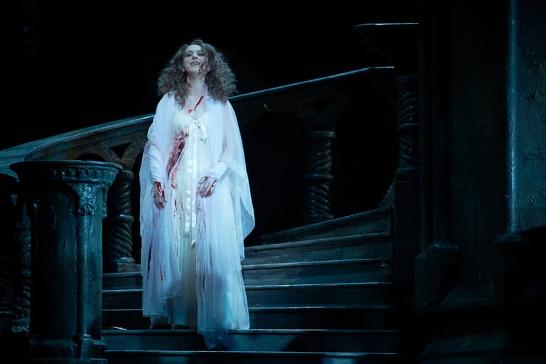 Performer in flowing white gown stands on dimly lit stage steps under blue theatrical lighting.